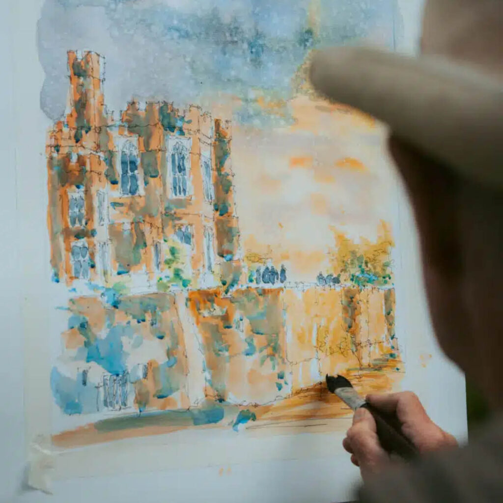 An unseen person paints a watercolour depiction of Raby Castle using blue and yellow paints.