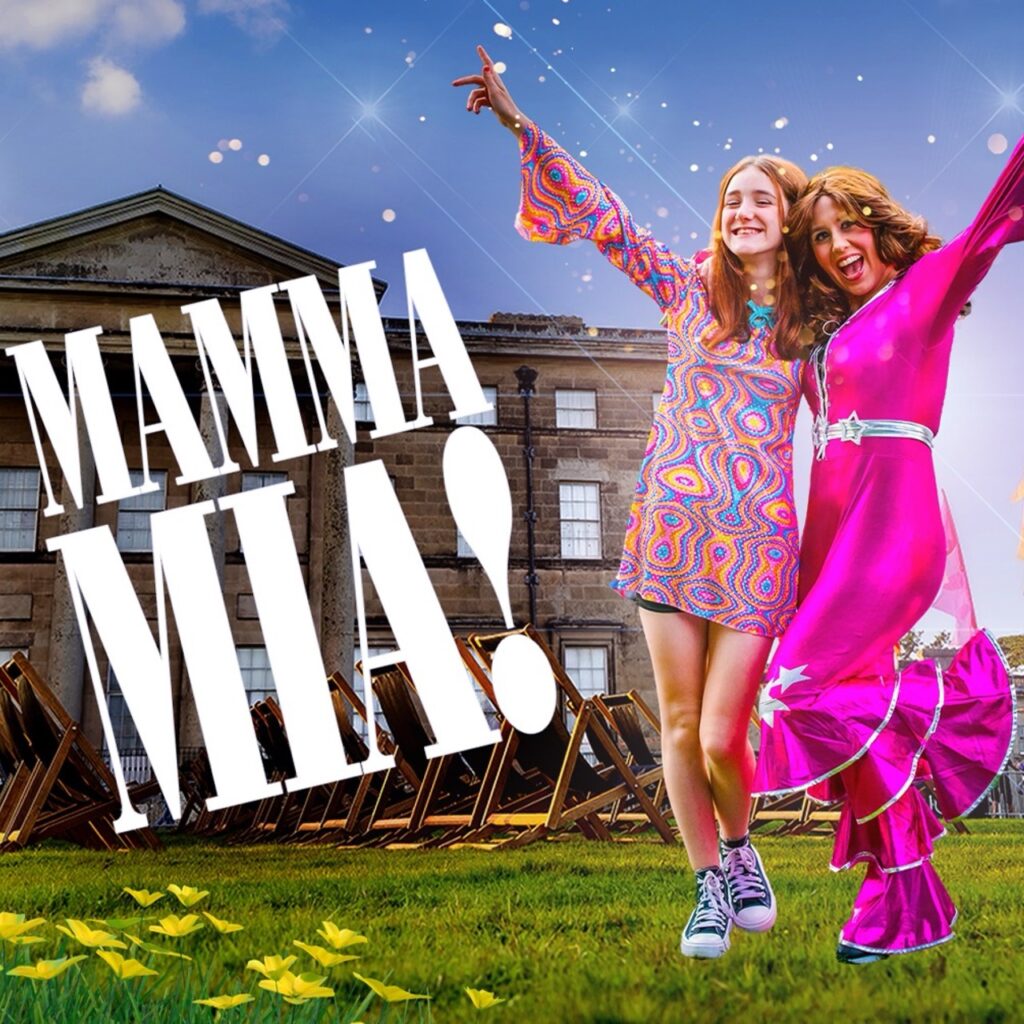 Two women dressed in pink disco style dresses raise their hands in the air as they sing along to an ABBA song. Big white words next to them say: Mamma Mia!