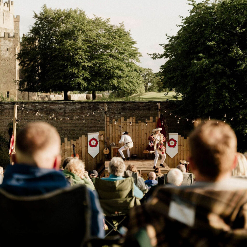 People sit on deck chairs in the grounds of Raby Castle. They watch a wooden stage where two performers dressed in Elizabethan clothes run around the stage.