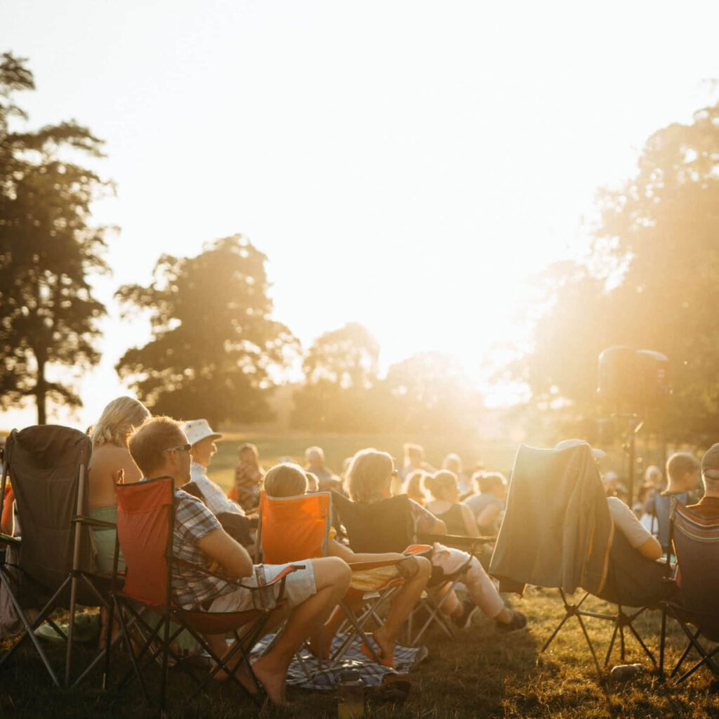 People sit on deckchairs in Raby Castle's grounds. The setting sun shines through the trees as the people watch an unseen cinema screen.