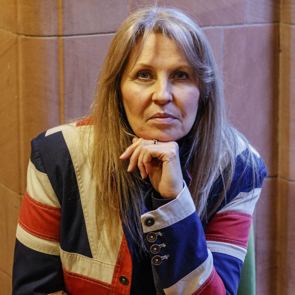Philippa Langley is a white woman with long blonde hair. She wears a jacket in union jack colours. She rests her chin on her hand and looks seriously at the camera.