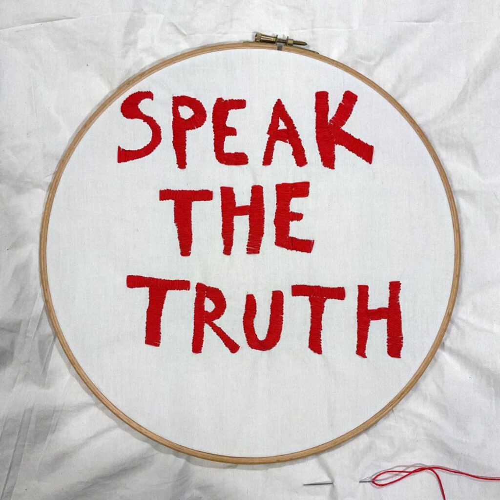 Red letters have been stitched onto a white embroidery hoop. They say Speak the Truth. This is part of Delaine Le Bas's performance work.