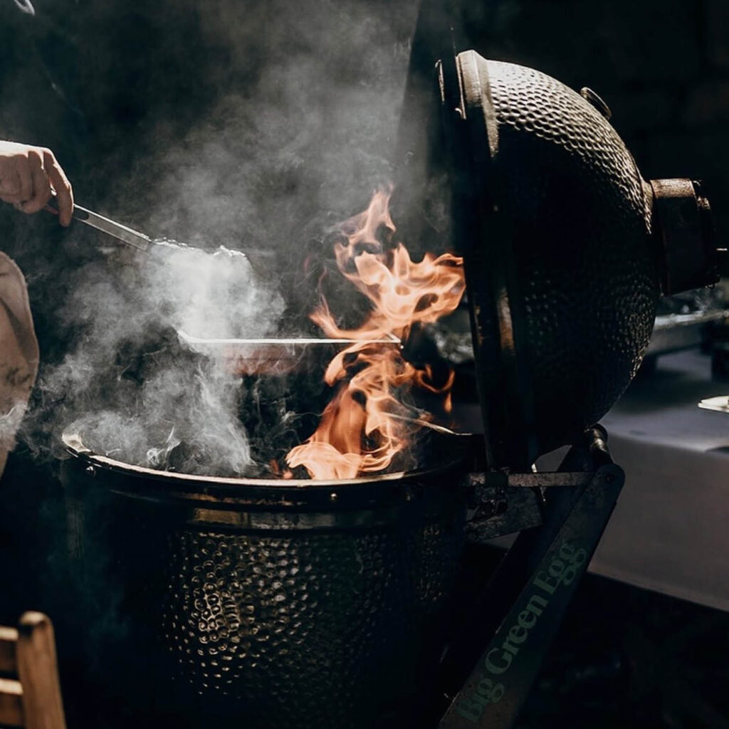 Flames rise from a BBQ grill as someone uses tongs to cook food.