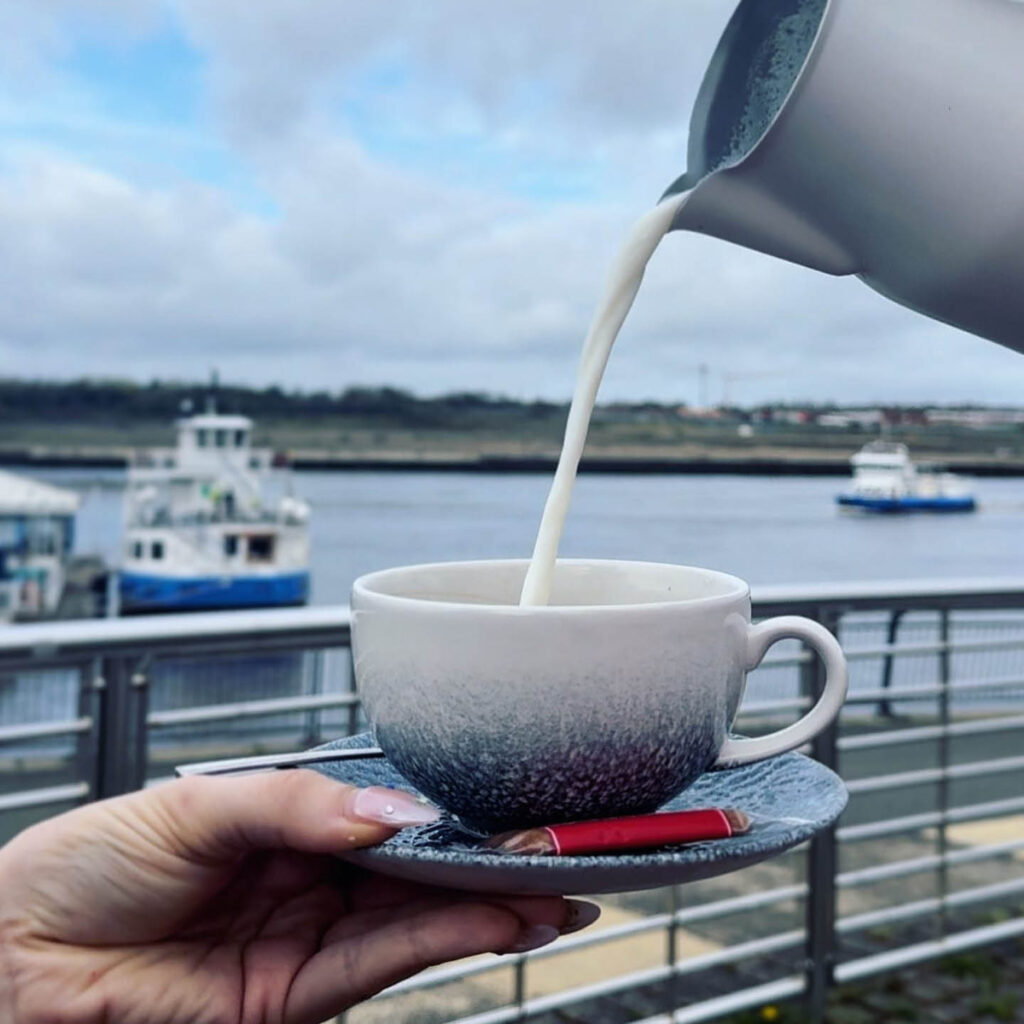 A hand holds up a blue and white teacup on a saucer. A milk jug is raised above it and pours a stream of milk into the cup. In the background, a couple of boats float in the South Shields harbour.