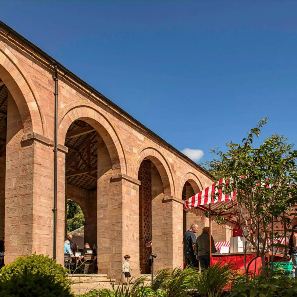 A large brick viaduct looms against a deep blue summer sky. Red and white striped market stalls are dotted beneath it. This is the Harvest Market at Raby Castle.