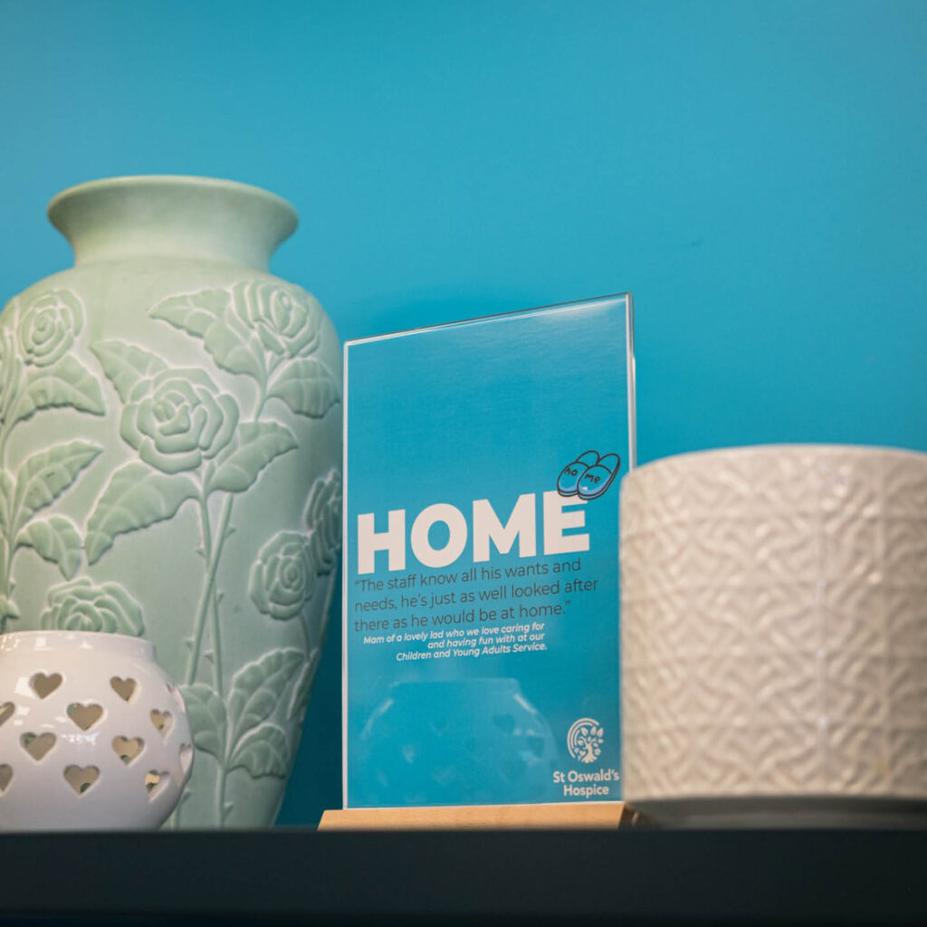 A green vase, a white plant pot and a white tea light holder are positioned on a shelf against a bright blue wall. At the centre of them, a blue sign displays a quote from a user of St Oswald's Hospice.