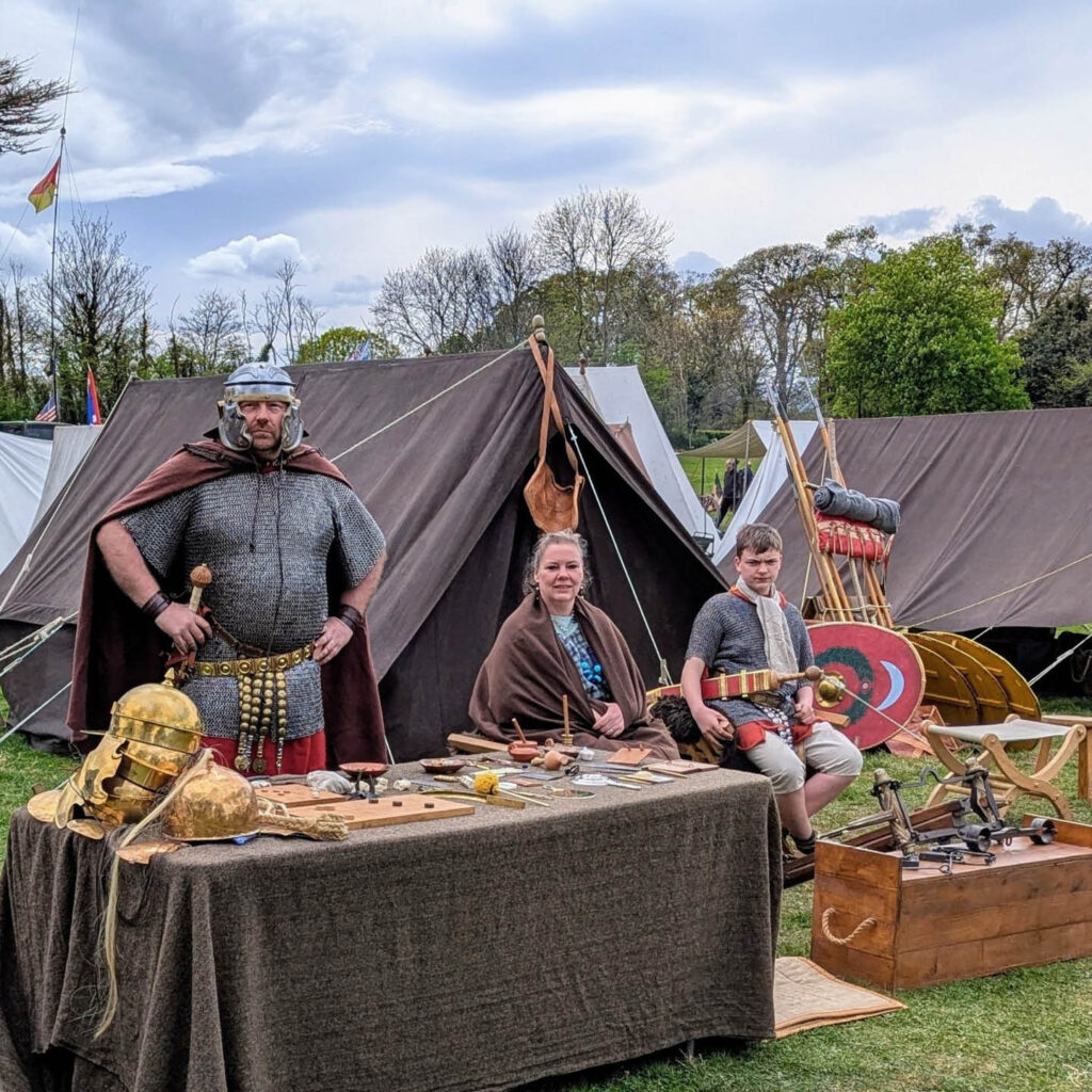 A fully armoured soldier stands behind a table displaying tools and arms. A woman wrapped in a shawl sits beside him, along with a boy carrying a sheathed sword. They are gathered outside of an old canvas tent and look at the camera. They are part of the reenactment group for the Meet the Tungrians event at Arbeia South Shields Roman Fort.