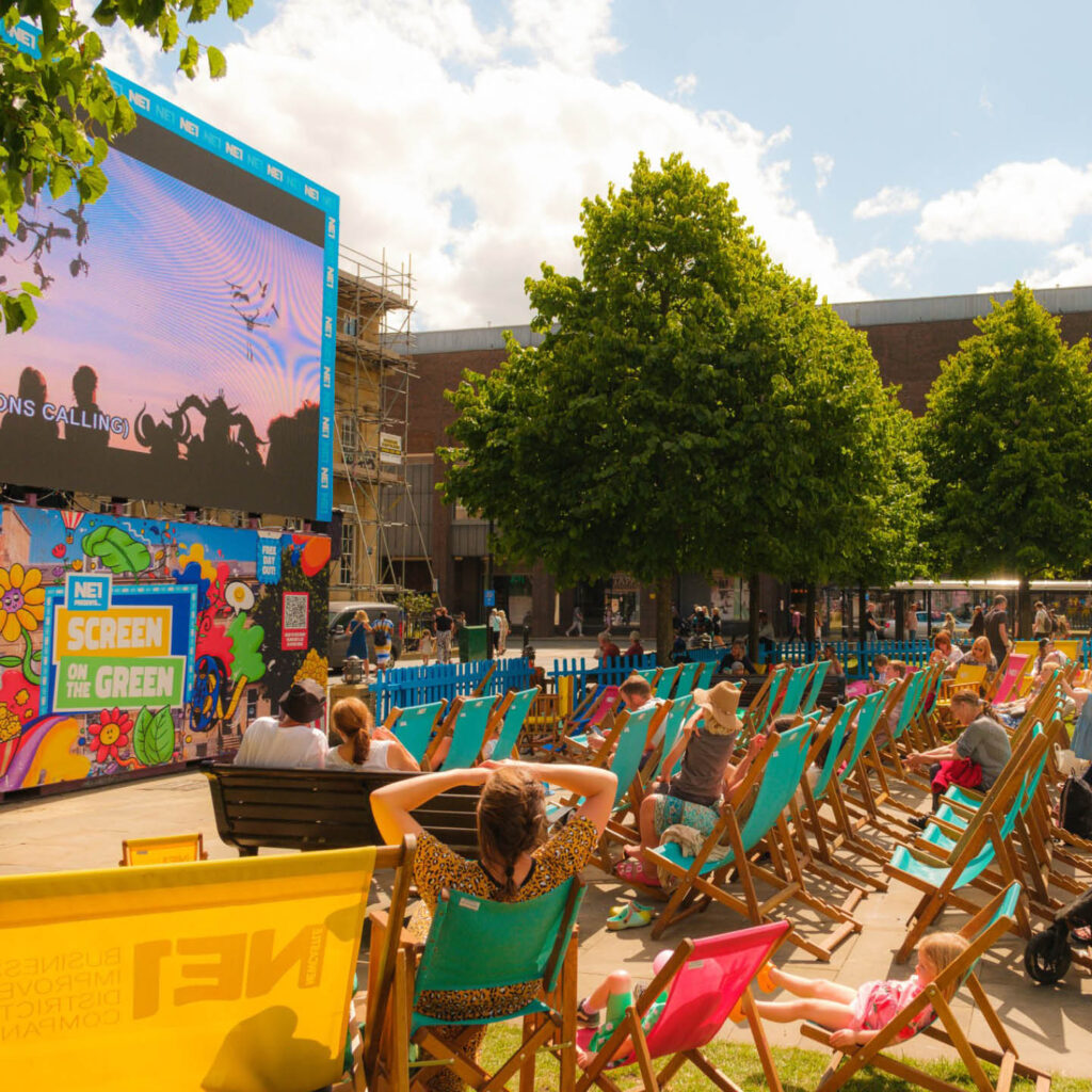 People lounge on blue and pink deckchairs arranged in front of a big open-air screen. The sun shines down. This is NE1's Screen on the Green in Old Eldon Square, Newcastle.