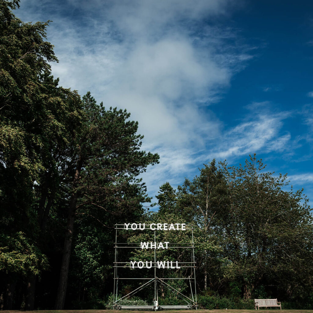 Illuminated text is fixed to a large scaffold. It says: You Create What You Will. Dark trees and bright blue sky loom behind it.