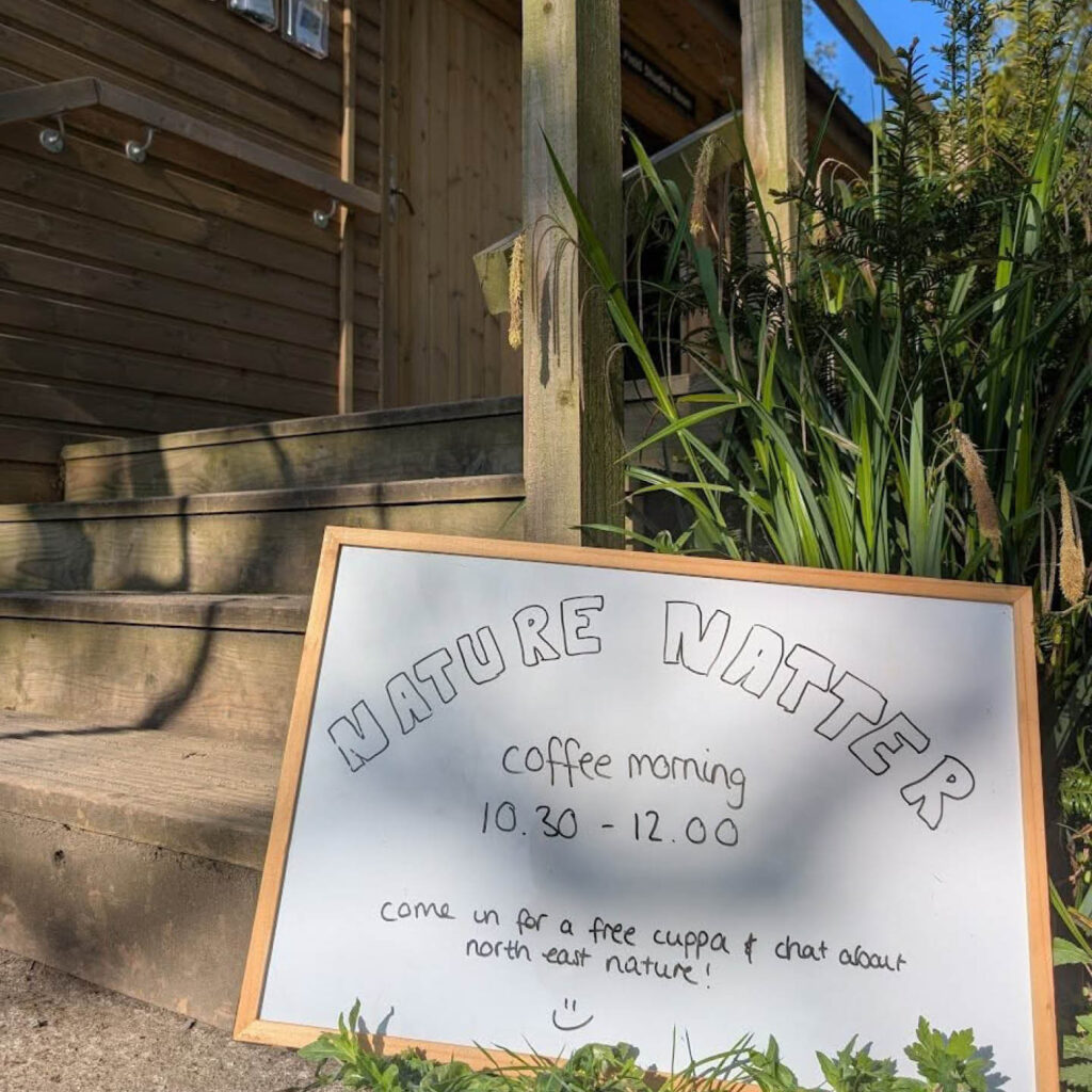 A small whiteboard sign sits in the grass outside a wooden building at Gosforth Nature Reserve. In black pen, someone has written: Nature Natter Coffee Morning, 10:30am to 12pm. Come in for a free cuppa and chat about North East nature.