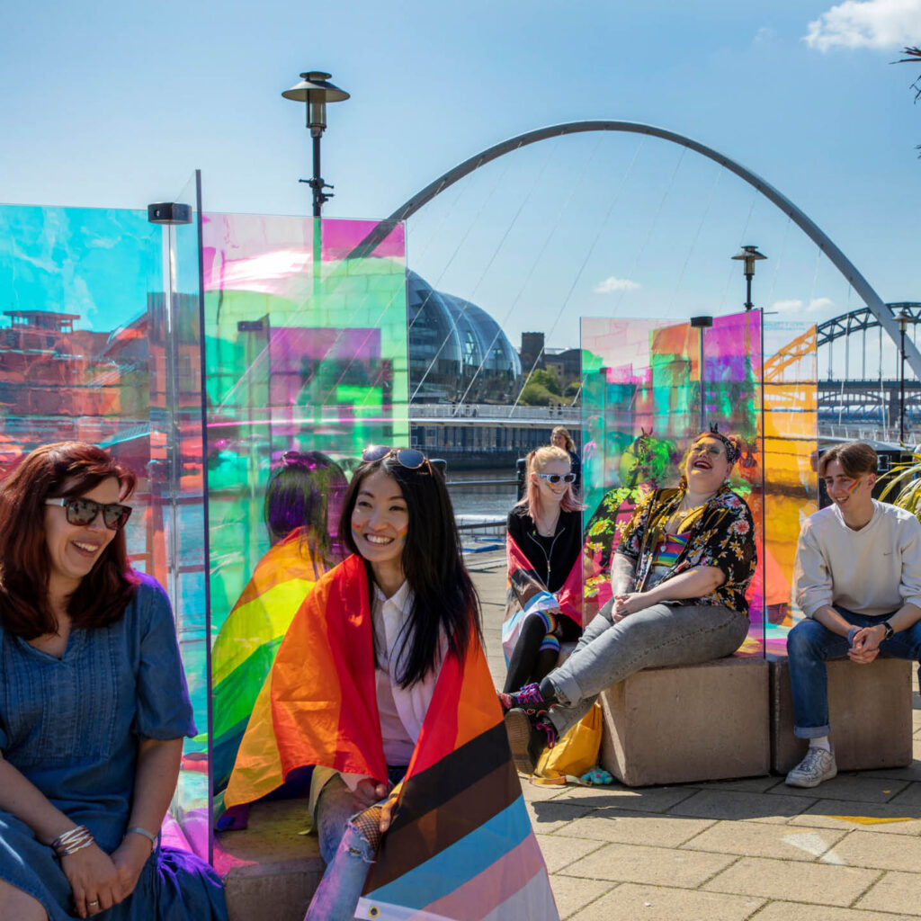 People sit on stone benches beside Newcastle's quayside. The sun shines down on them and reflects of multi-coloured glass partitions on the benches. The people wear rainbow flags and face paints.