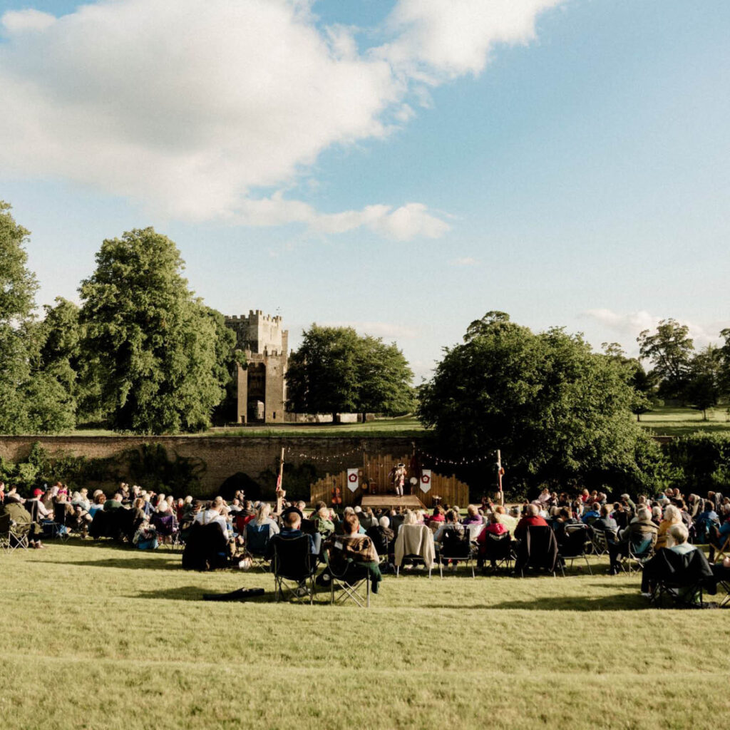 People are seated on deckchairs at the centre of a grass amphitheatre at Raby Castle. They watch a performance on a wooden stage. The Castle can be seen in the background surrounded by trees beneath a blue sky.
