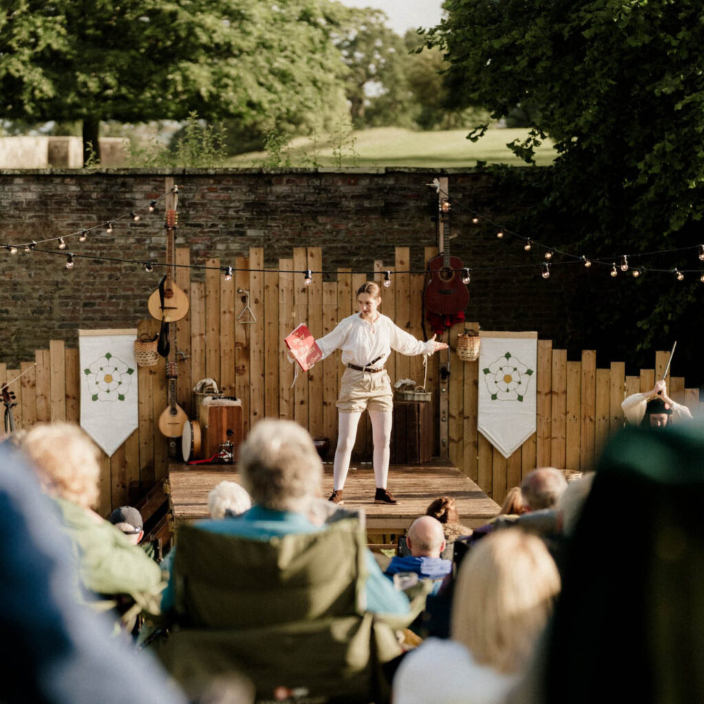 A person dressed in white tights, puffy beige shorts and a white blouse gestures with both arms as they stand on a wooden stage at Raby Castle. People watch the play as they sit on deckchairs in the sun.