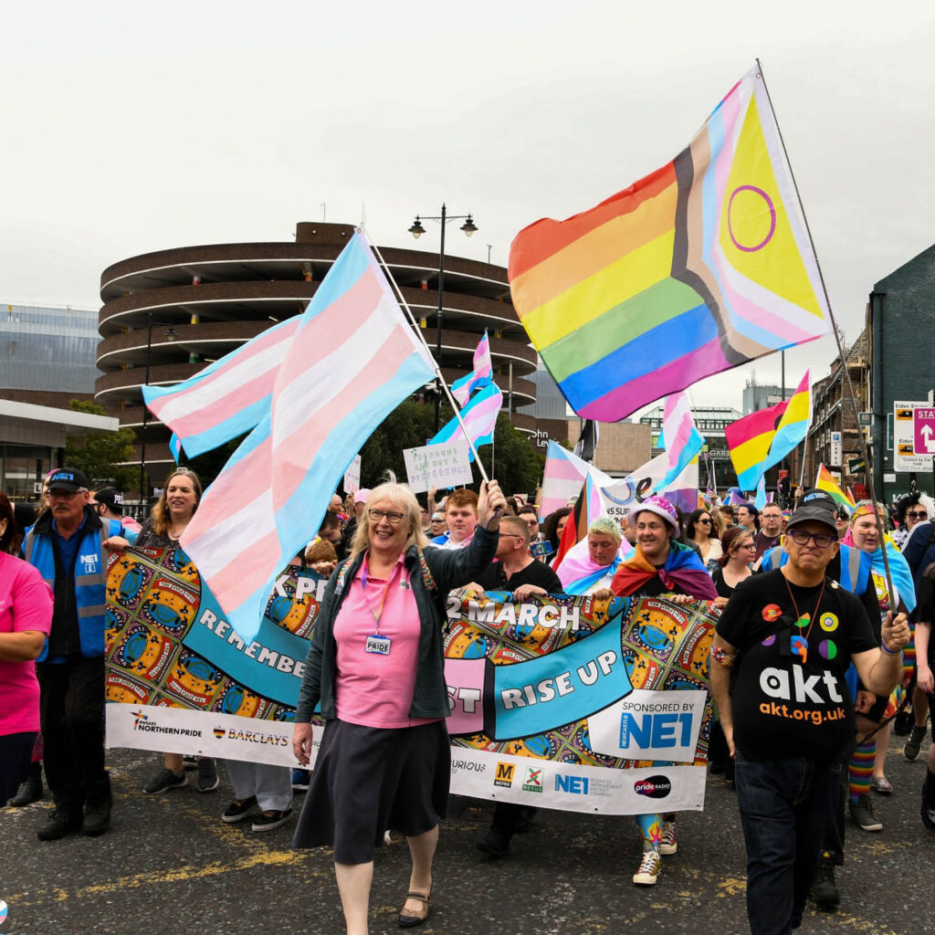 People smile and march down a street in Newcastle carrying banners and flags with the LGBTQIA+ colours.