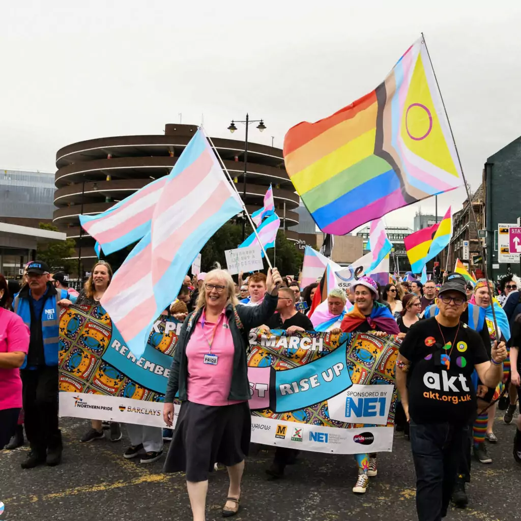 People smile and march down a street in Newcastle carrying banners and flags with the LGBTQIA+ colours.