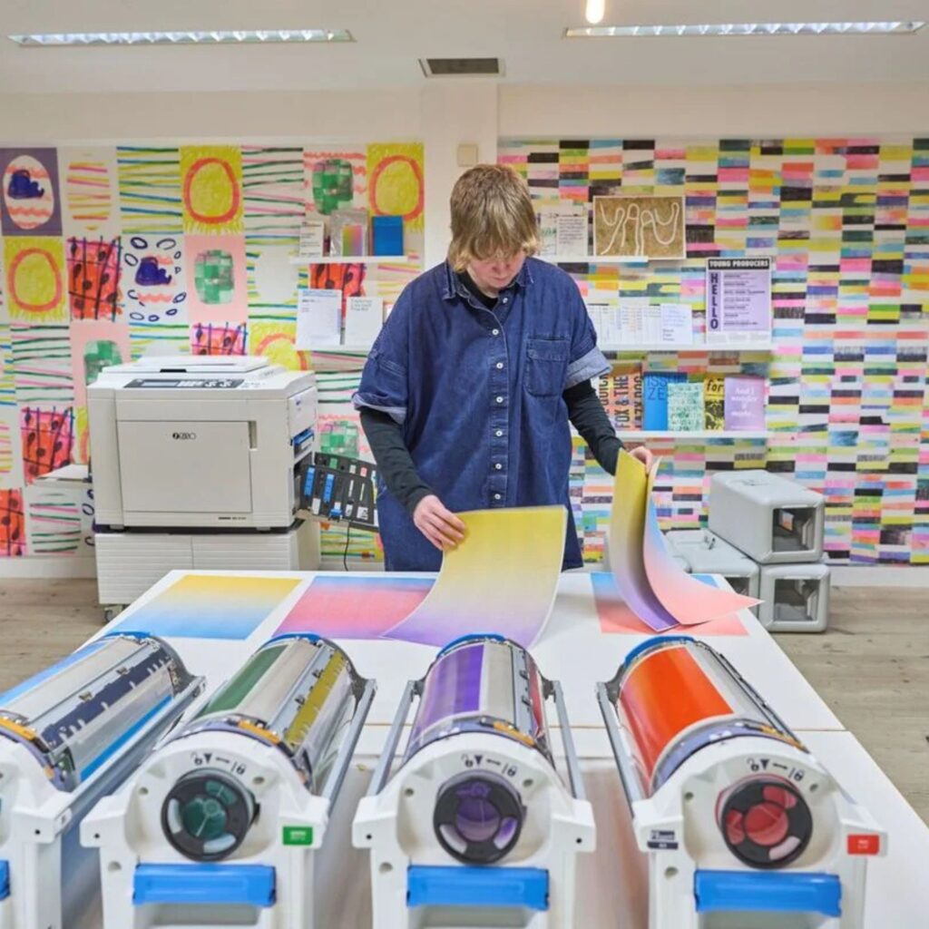 A person flips through coloured sheets of paper. They stand in front of cylinders of ink in one of the studios at the BALTIC. They are leading one of the risograph printing workshops.