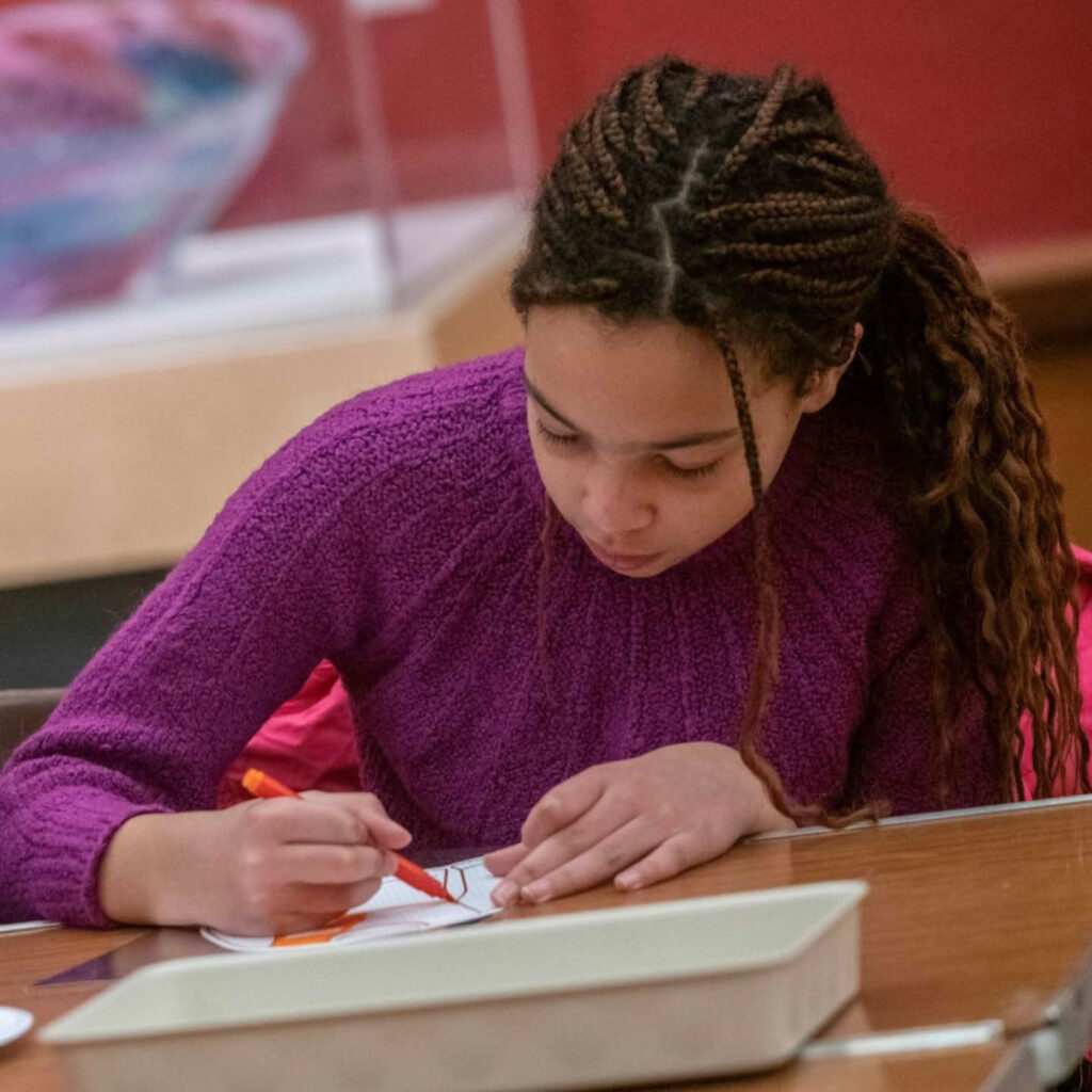 A young woman in a purple jumper sits at a table in the Laing Art Gallery. She uses a red pen to draw on a piece of paper.