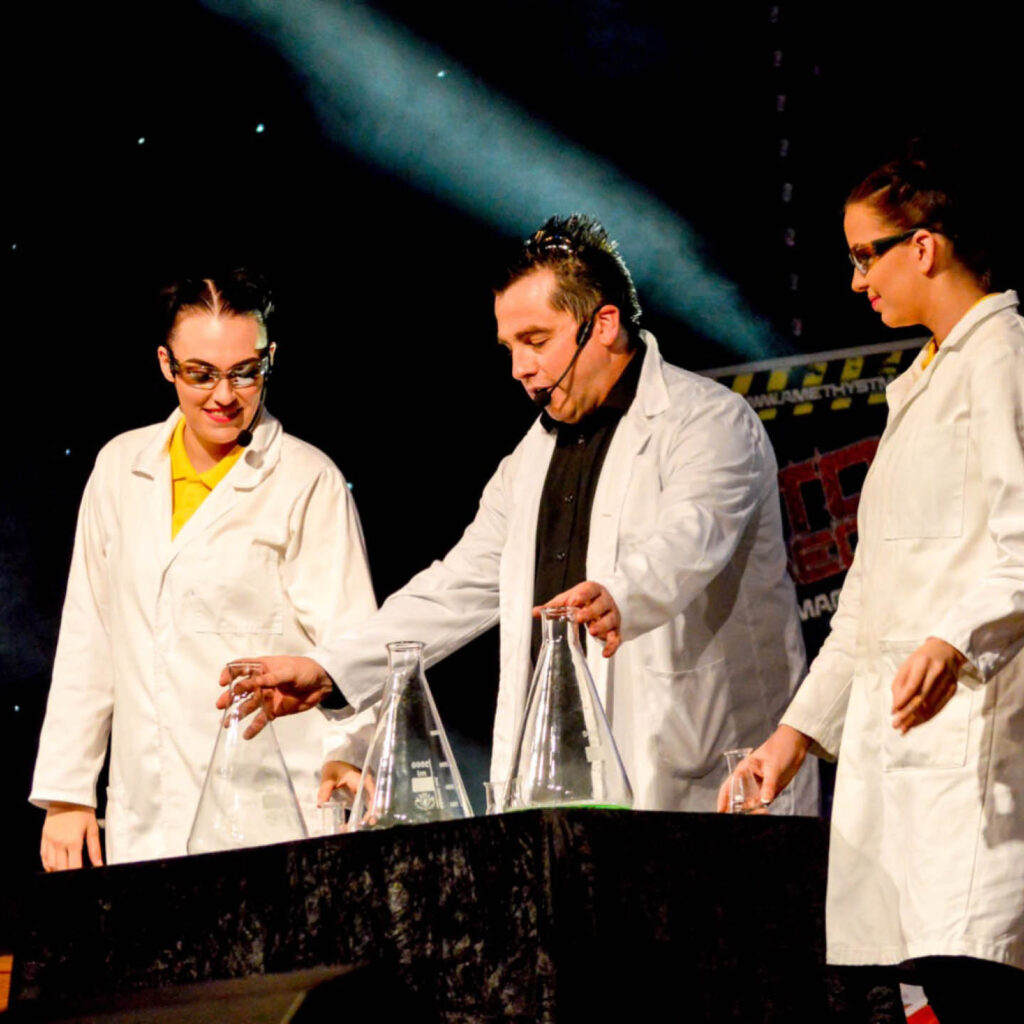 Three scientists in lab coats and goggles grin as they look down at three conical flasks on a black table. They are part of the stage show: The Magic of Science.
