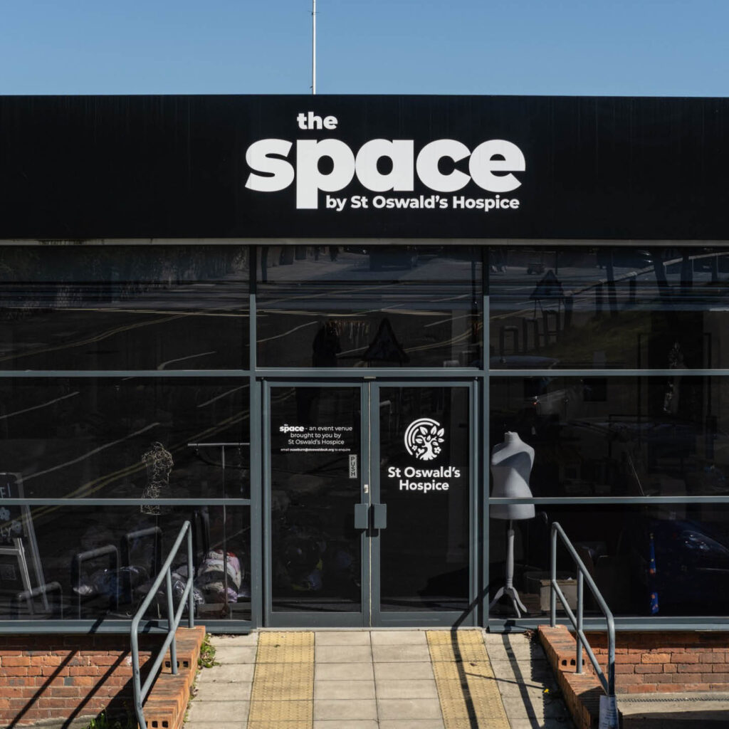 Glass store front with a black sign above the door. White lettering says: The Space by St Oswalds Hospice.