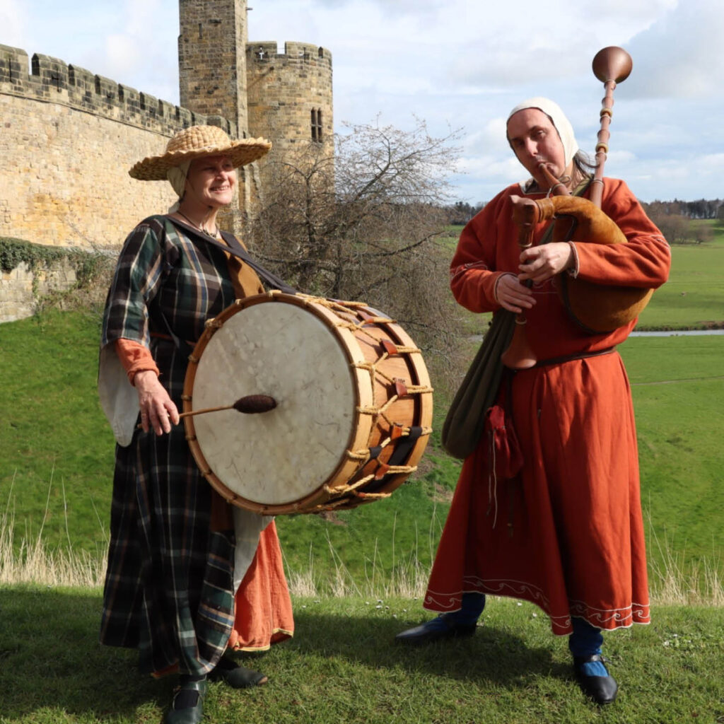 A man and woman dressed in medieval hats and tunics stand in the sunny grounds of Alnwick Castle. The woman smiles as she hits a large drum strapped to her front. The man blows into a bagpipe. They are part of the ensemble Trouvère, who play medieval music at Alnwick Castle.