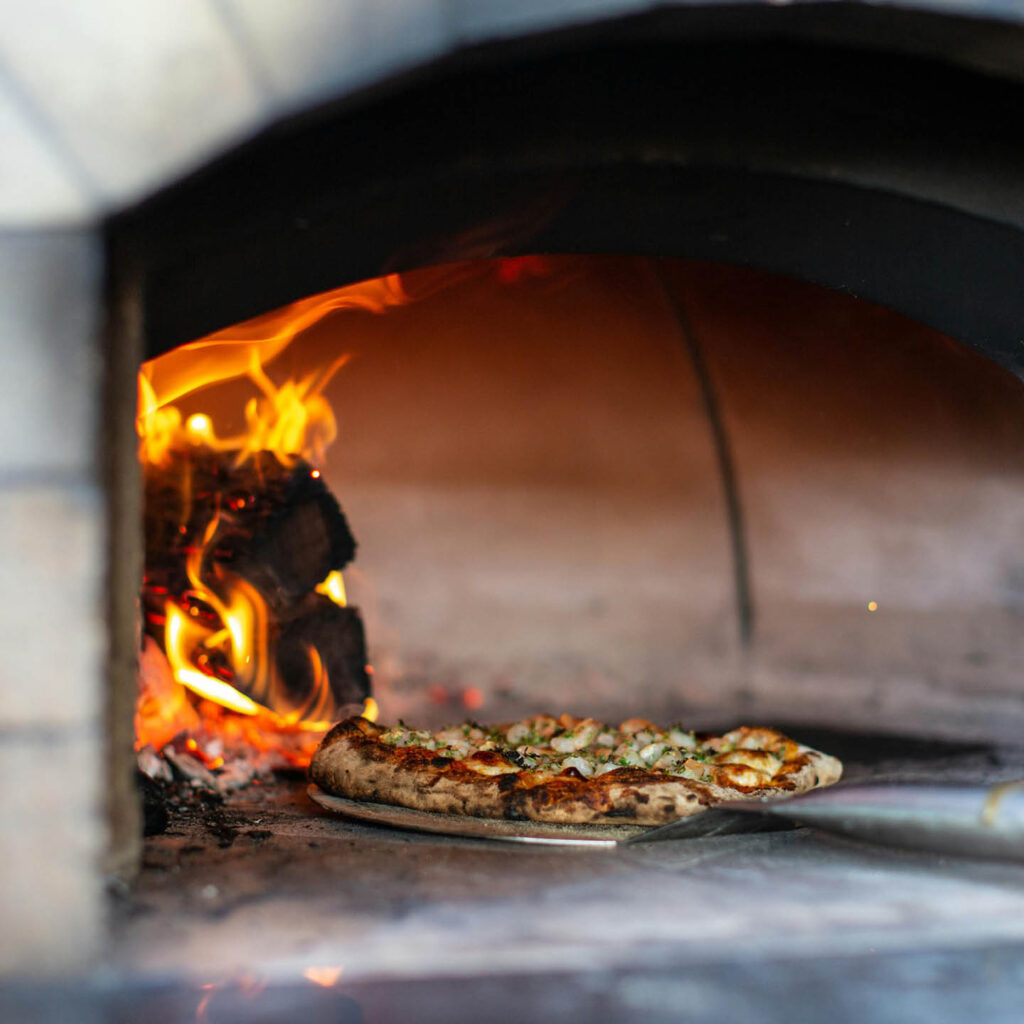 A pizza is placed inside a wood fired oven using a metal pizza peel.