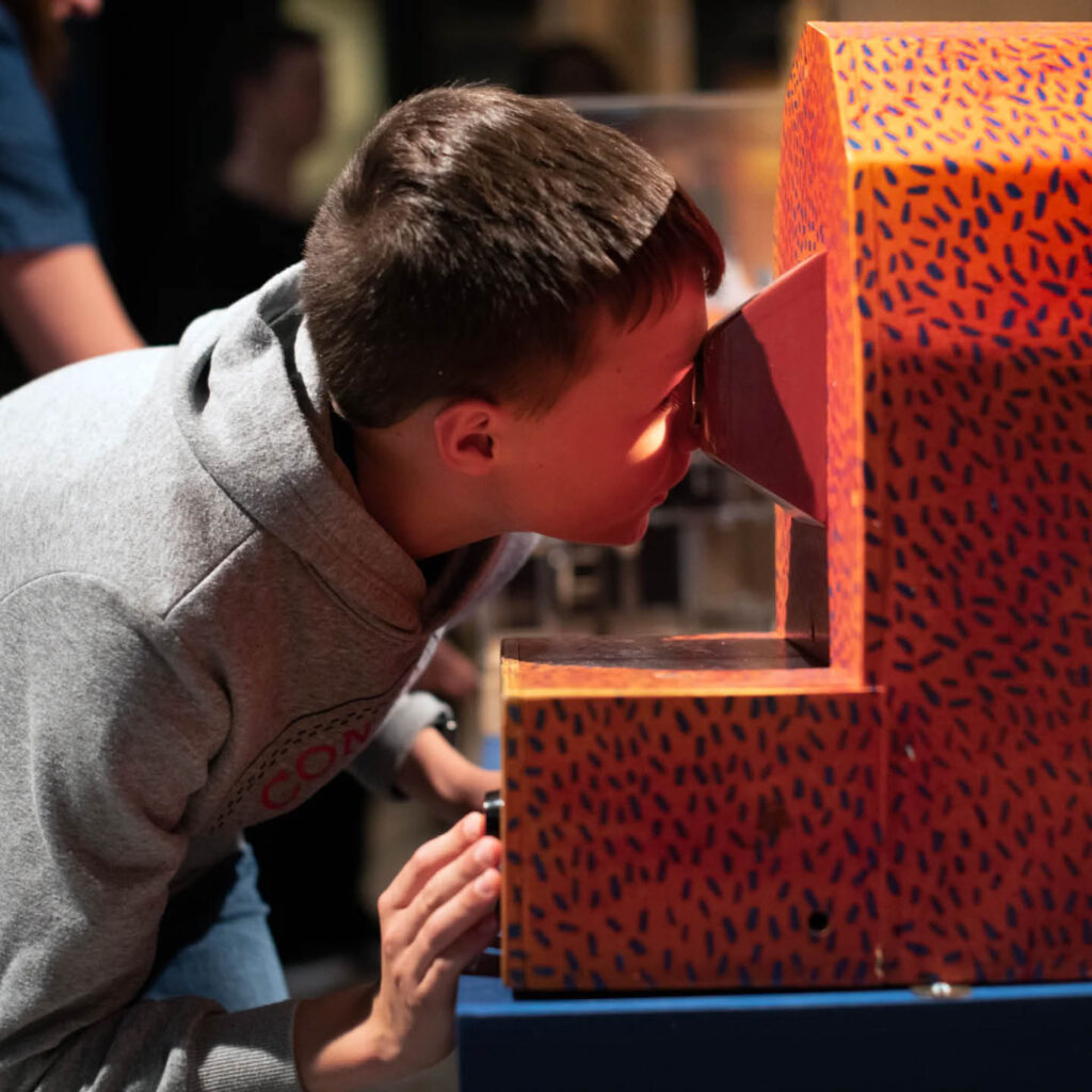 A young boy crouches down to peer inside an orange periscope looking contraption. He is taking part in an Arty Steam session at The Bowes Museum.