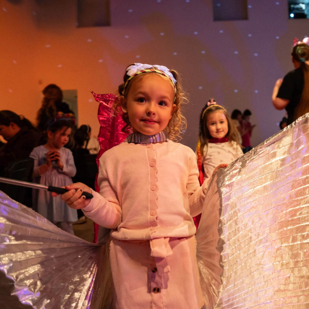 A little girl dressed in pink smiles as she holds open shiny silver wings attached to her back. She stands in a dimly lit room with white disco lights reflected on the wall behind her. She is taking part in one of the BALTIC's Big Summer activities.