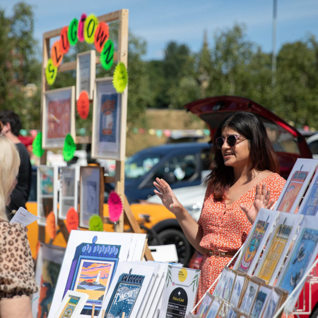 A woman in a pink dress and sunglasses stands behind her stall where she is selling prints. She gestures as she chats to visitors browsing her art.