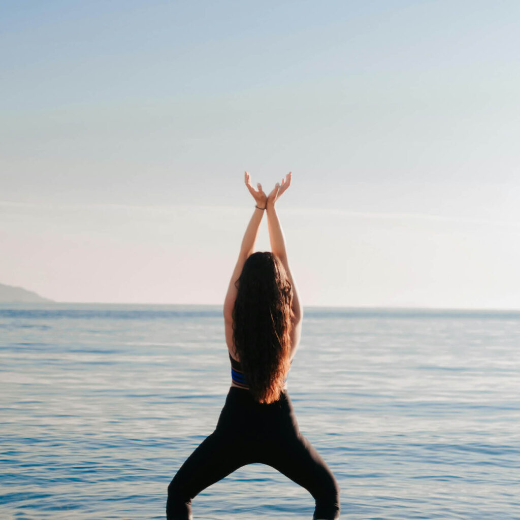 A woman in black sports clothes and long brown hair raises her arms above her head. She looks out across the sea.