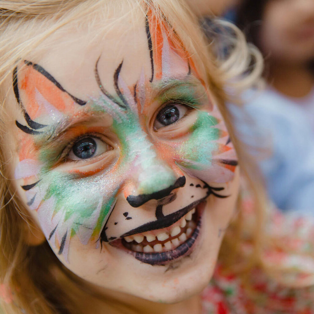 A little girl grins up at the camera. Her face is painted like a ginger and green cat.