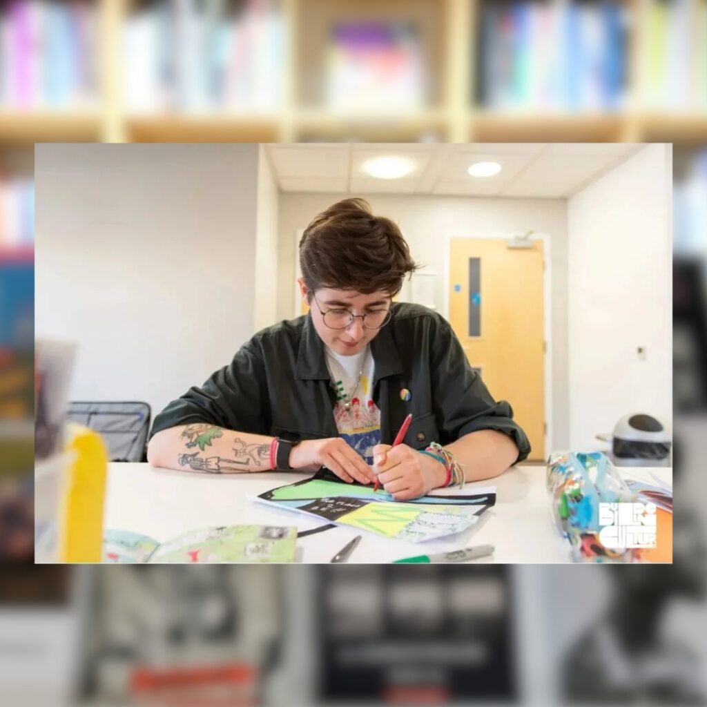 Artist Mack Sproates sits at a table inside the BALTIC and draws on a sheet of paper. They are leading the Climate Home Zine workshop at the BALTIC.