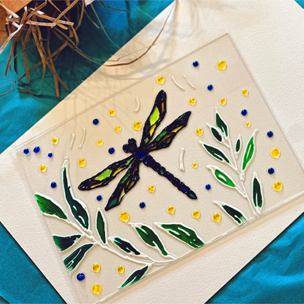 A glass rectangle with a colourful resin embellishment of a dragonfly and plants.