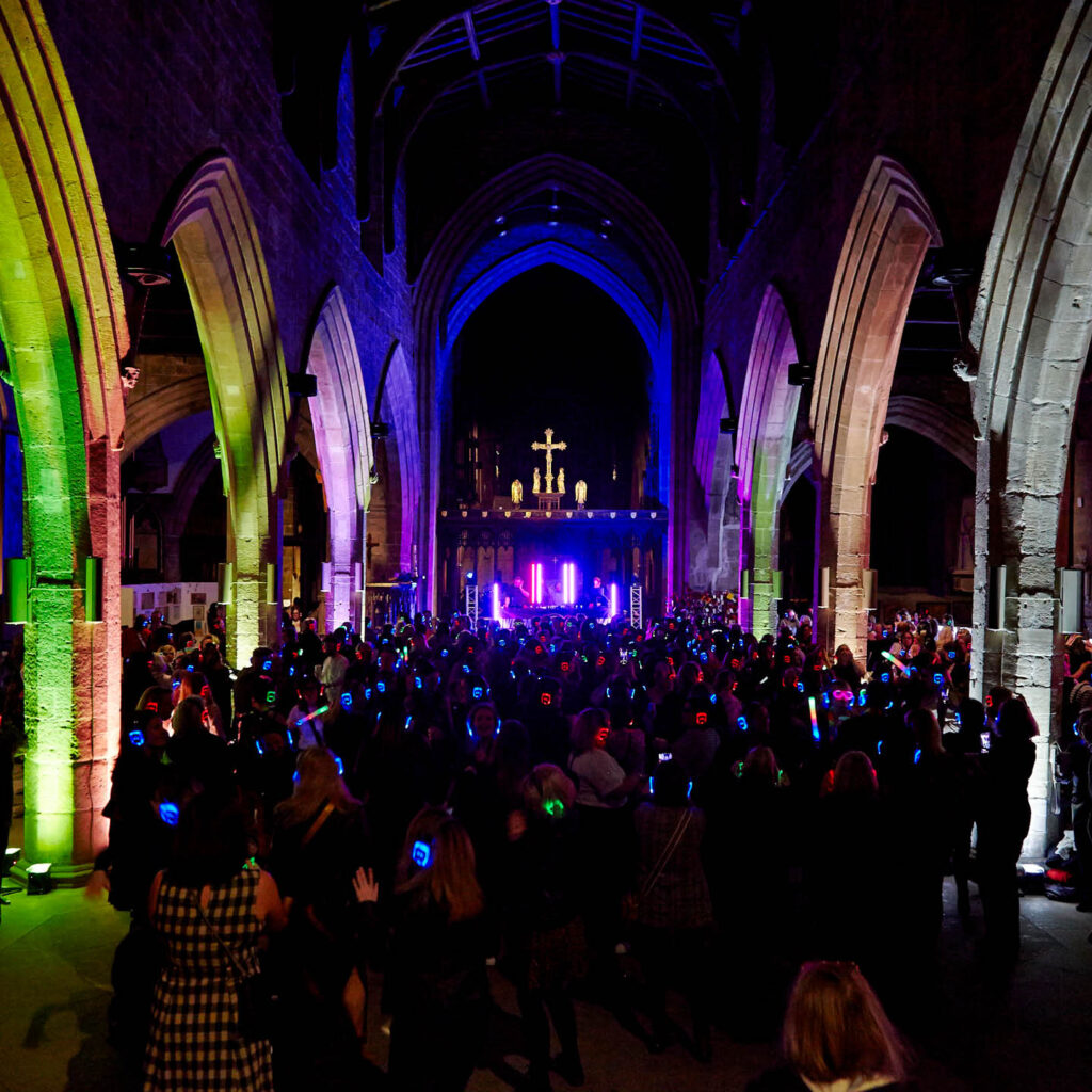 The stone arches in Hexham Abbey are illuminated with purple, green and pink light. Hundreds of people fill the dark space wearing light-up headphones. They are enjoying the Awesome Silent Disco event at the abbey.