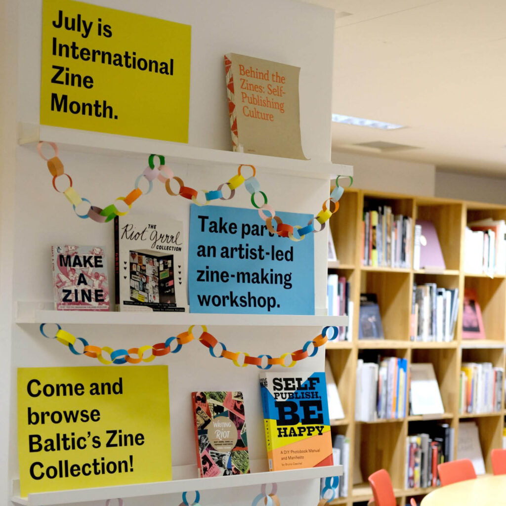 White shelving inside the BALTIC's library is strung with colourful paper chains. Yellow and blue posters explain that July is International Zine Month and invite people to browse the zine collection and take part in artist-led workshops. A collection of zines are placed aesthetically on the shelves next to these posters.