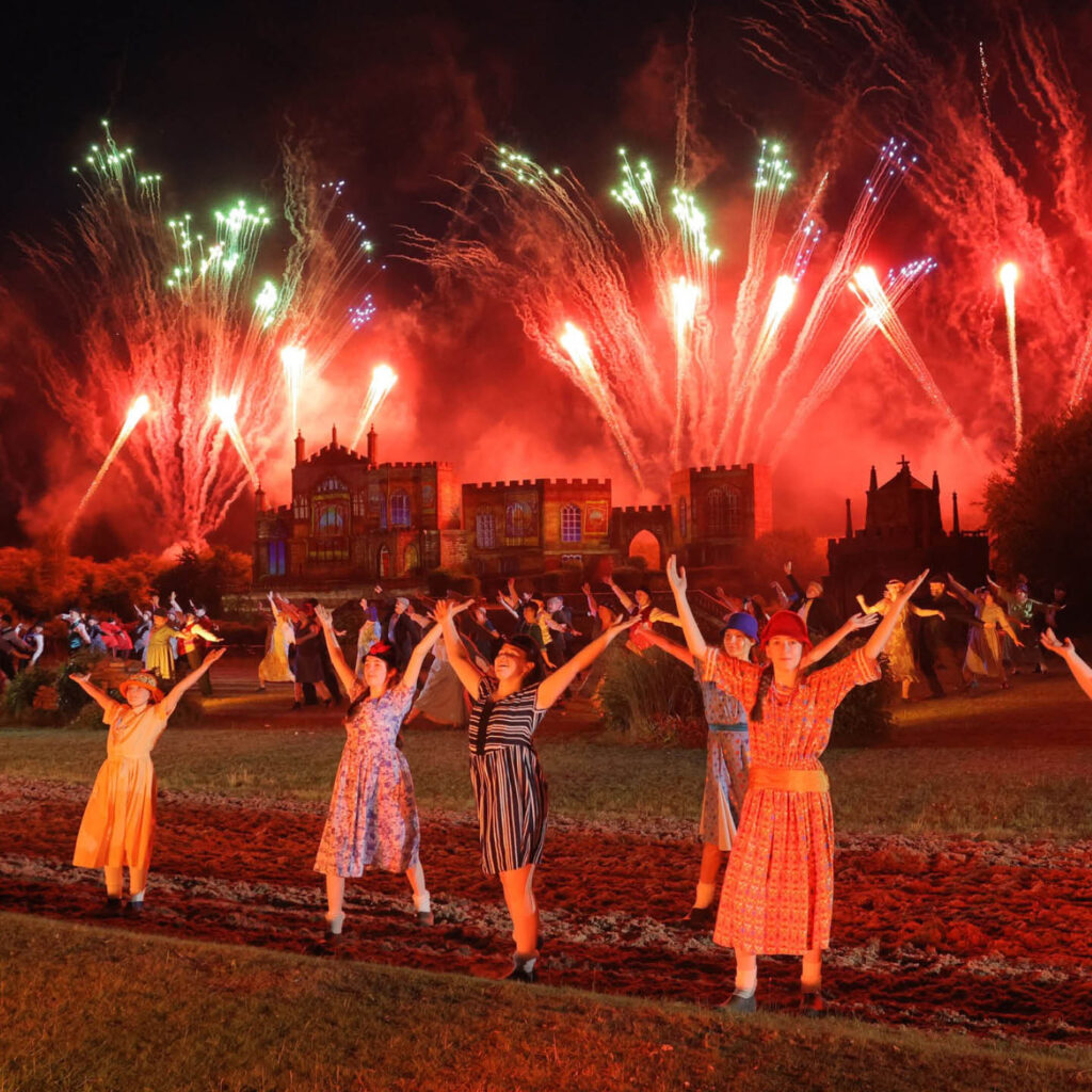 Women dressed in floral dresses and hats raise their arms in the air as a huge display of red fireworks bursts behind them above Auckland Palace. They are part of the annual Kynren show.