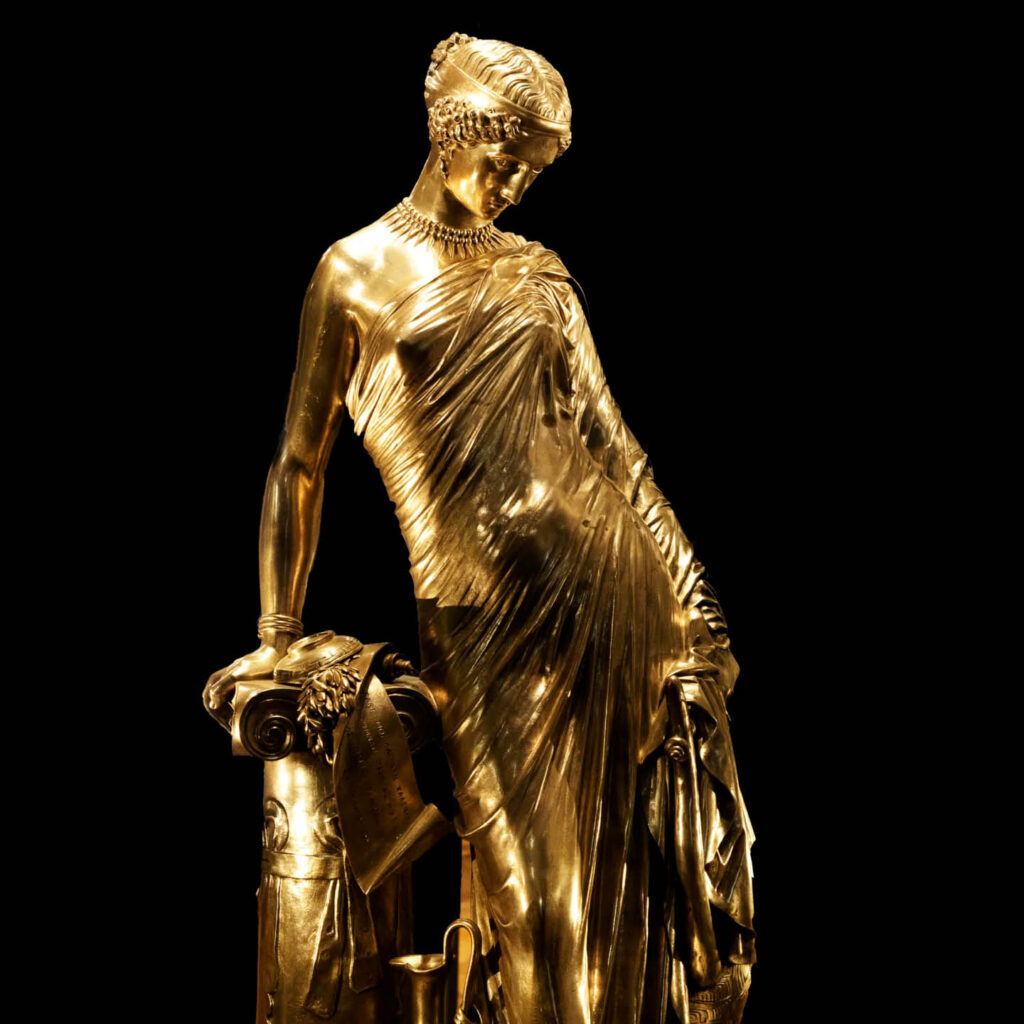 A gold statue of a woman from ancient Greece. Her body is covered by a silken looking shroud and she leans one arm on a rock behind her.