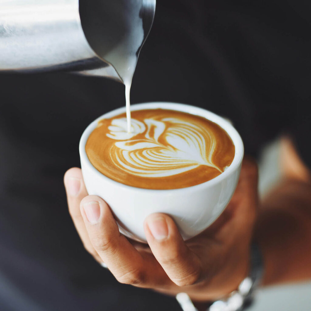 A person skilfully pours milk into a cup of coffee, creating a beautiful flower on top of the foam.
