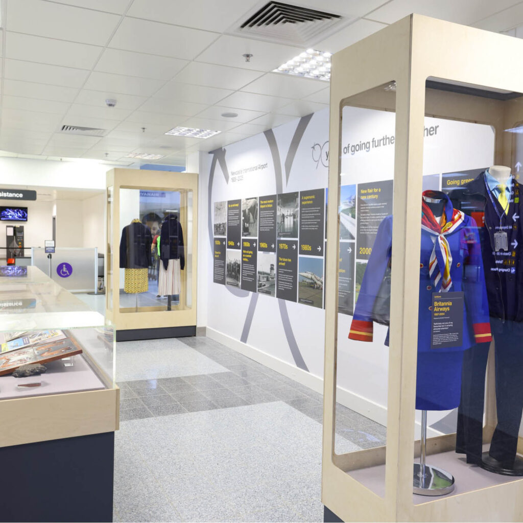A display case holding two vintage cabin crew uniforms. A wall displays information about Newcastle Airport's 90-year history. More cabinets and display cases can be seen in the background.