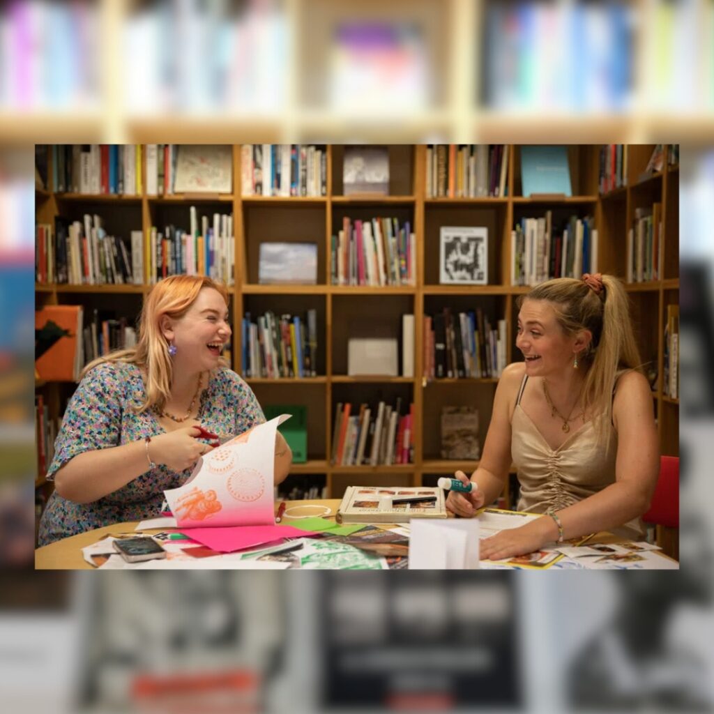 Two women laugh as they create zines together in a room at the BALTIC. Art books line a wall of bookshelves behind them.