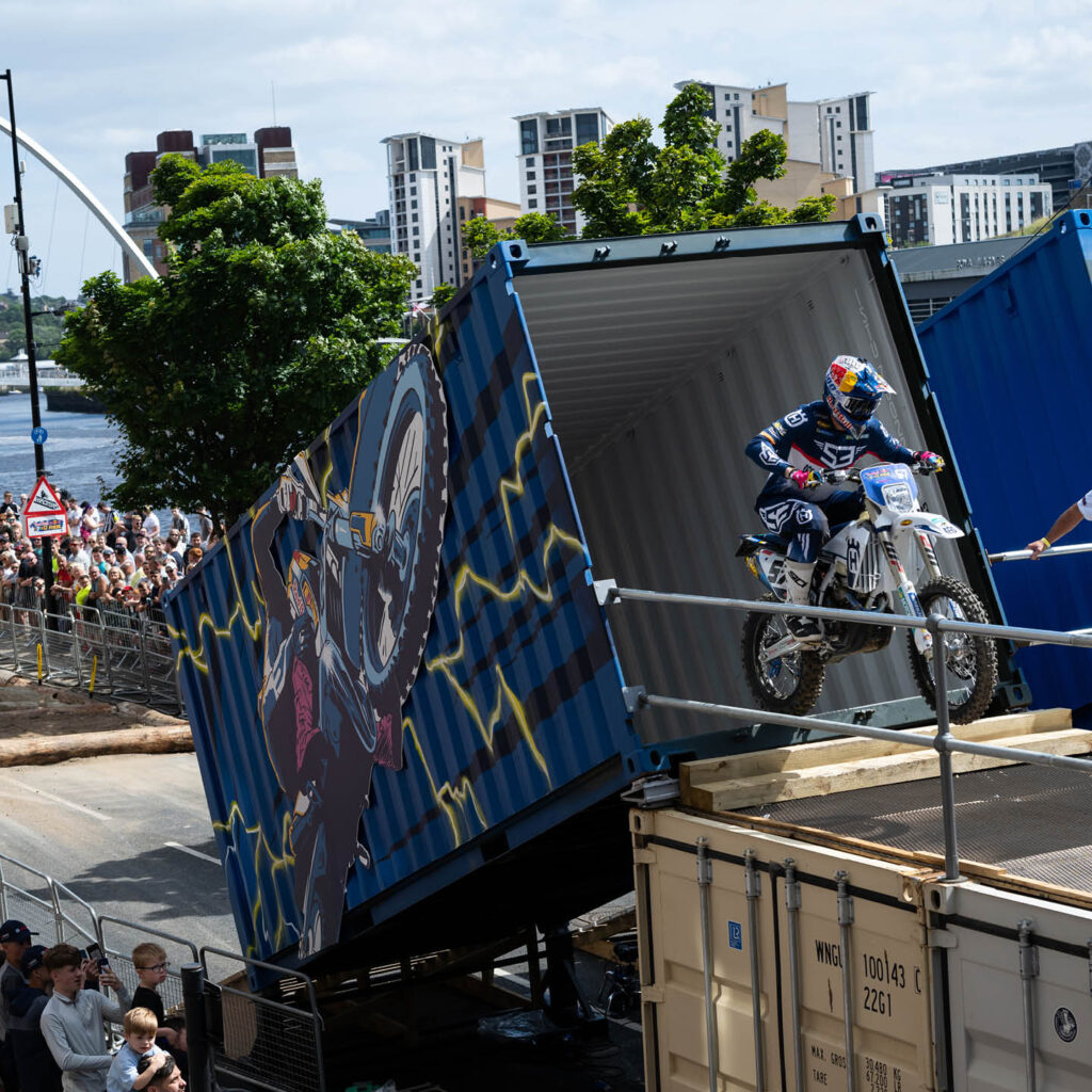 A motorbike rider comes out of the top of an open ended shipping container which has been propped up to create a ramp. Crowds gather in the background to watch the Red Bull Tyne Ride.