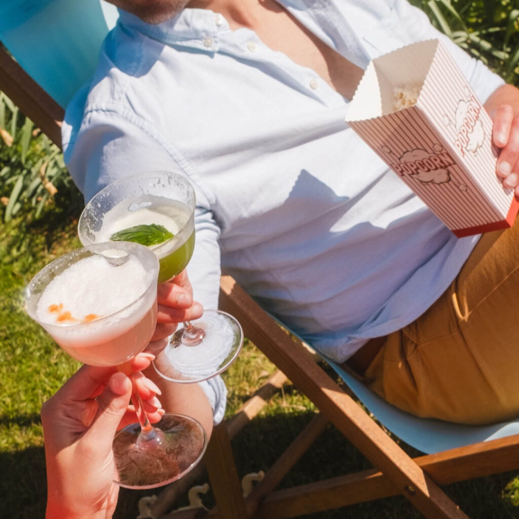 Two people clink cocktail glasses in the sun at Rockliffe Hall. One of them is seated in a deck chair and holds a paper tub of popcorn.