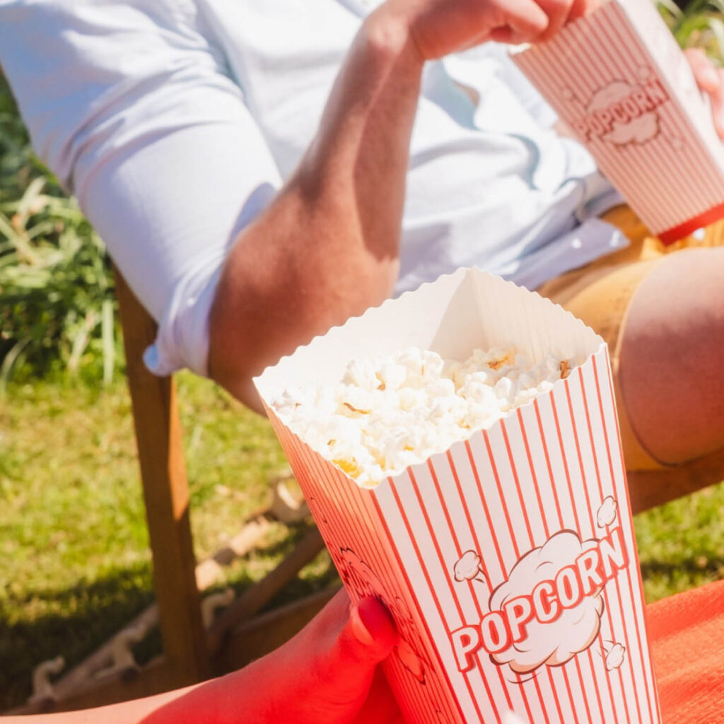 Two people hold paper tubs of popcorn in the sun. They sit on deck chairs on the sunny lawn at Rockliffe Hall.