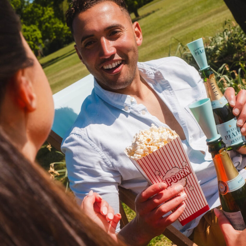 A man laughs as he holds a mini bottle of champagne and a box of popcorn. He talks to a woman in front of him who also holds champagne. The vibrant green grass of Rockliffe Hall can be seen behind them.