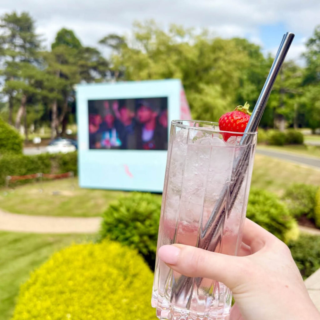 A manicured hand holds up a pink cocktail in a tall tumbler glass. In the background, a big cinema screen sits in the middle of a landscaped lawn at Rockliffe Hall.