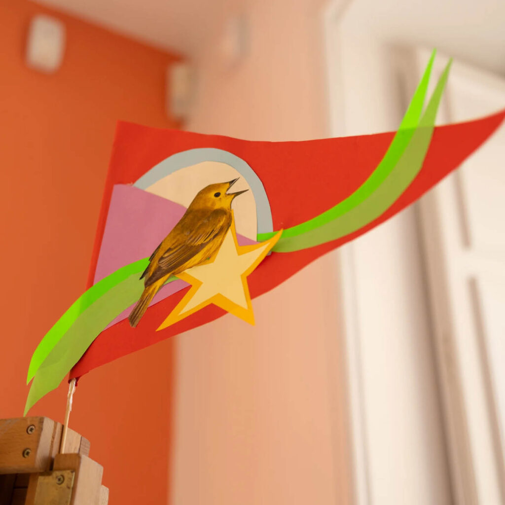 A red paper flag featuring a collage of a brown bird, star and green swirl. It is a piece of bunting from the Seaside Bunting workshop at The Bowes Museum.