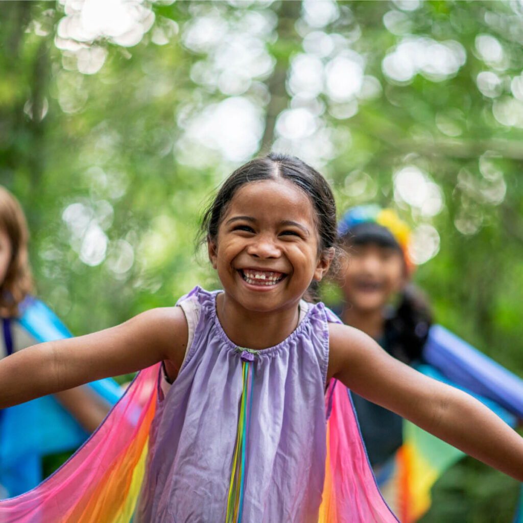 A little girl scrunches her face up in happiness as she runs through the woodland at Washington Wetland Centre. She wears a flowing pink cape which flies behind her.