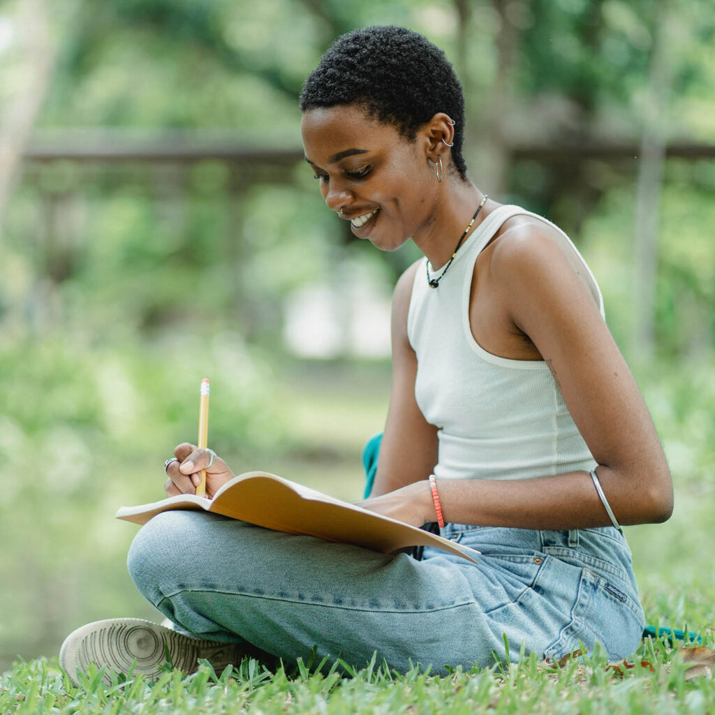 A young woman in jeans and a white vest sits cross legged on grass. She draws in the open sketchbook on her lap, smiling.