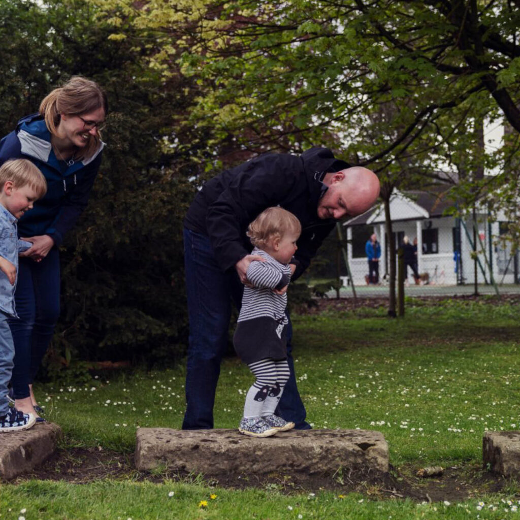 A dad crouches down to help his toddling son walk across a stone in the grounds of The Bowes Museum. Behind them, the mum does the same to a slightly older boy, who grins happily.