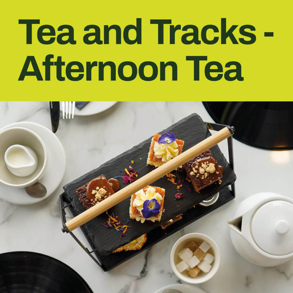 Pretty cakes and sponges are arranged on a black slate. Cubes of sugar and a pot of tea sit next to it. At the top of the image, black text on a green background says: Tea and Tracks, Afternoon Tea.