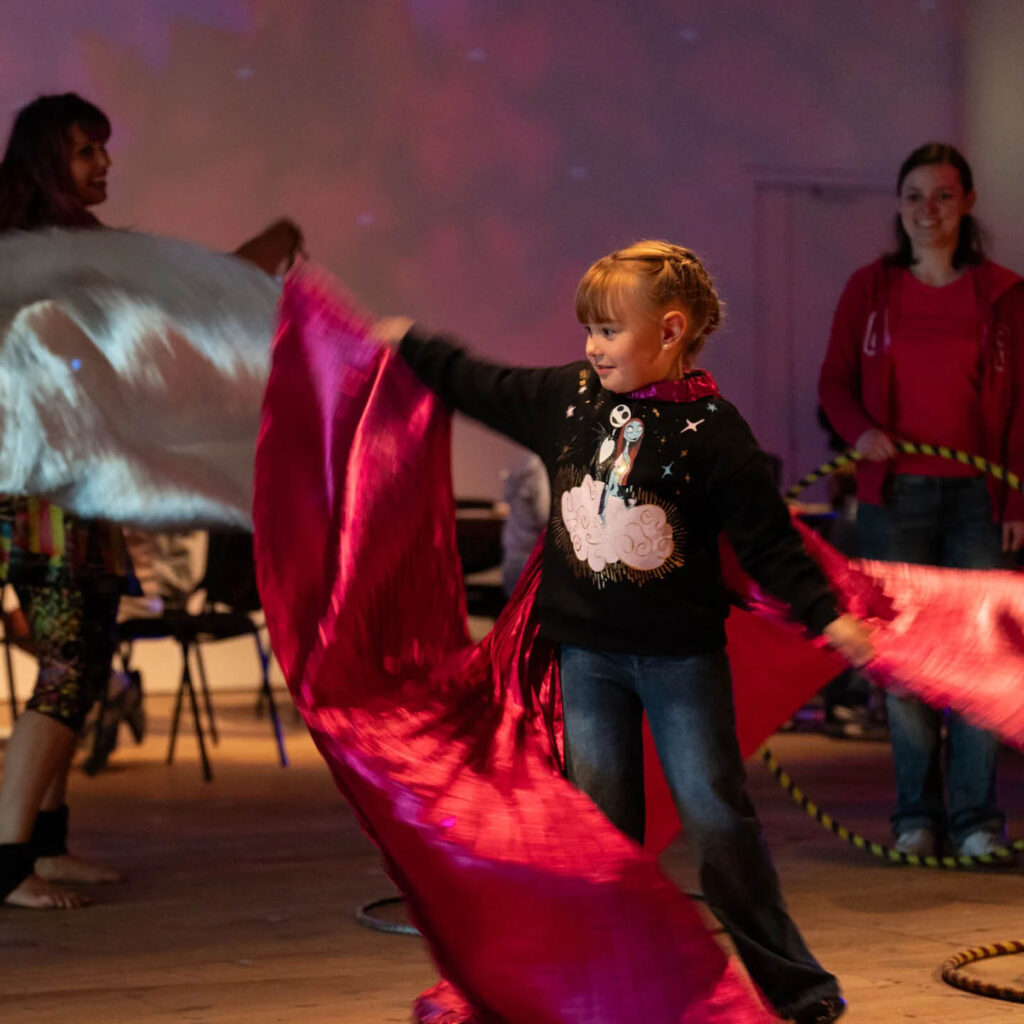 A little girl waves a red silky piece of fabric through the air. People smile and swirl behind her. The room is dimly lit with disco lights projected onto the wall. They are taking part in the Big Summer Disco at BALTIC.